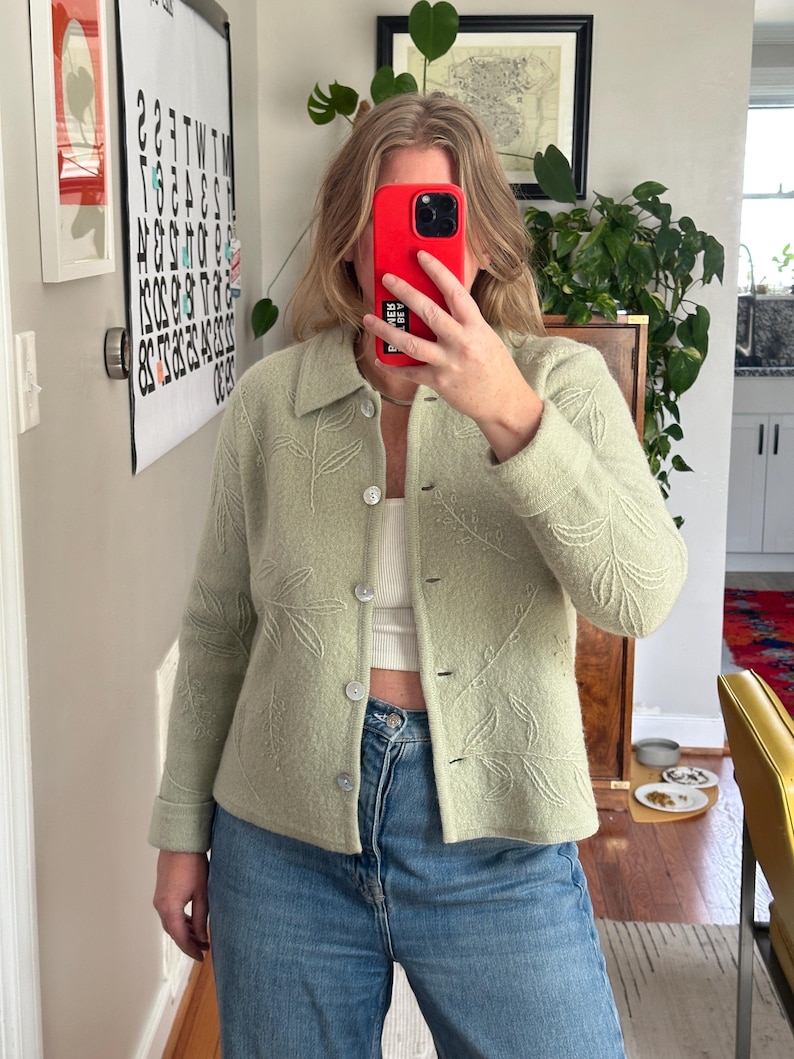 May include: A light green button-up jacket with a leaf pattern. The jacket has a collar and is worn over a white top and blue jeans. The jacket is made of a soft material.