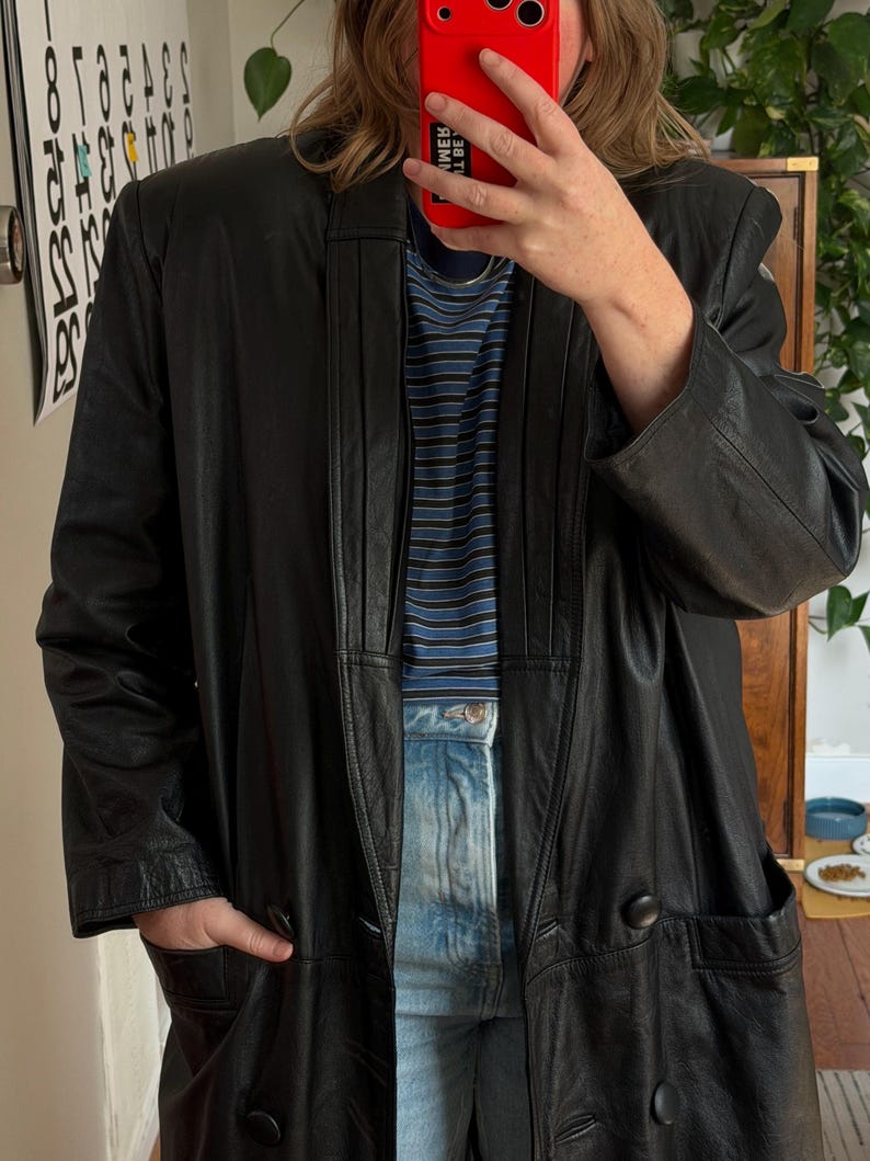 May include: A long black leather coat with a notched collar and two large buttons. The coat has two front pockets and a tailored fit. The person is wearing a blue and white striped shirt and blue jeans.