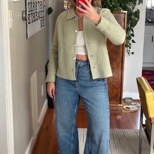 May include: A light green jacket with a leaf pattern, paired with blue jeans. The jacket has a button-up front and a collar. The jeans are a light wash, wide-leg style. The outfit is completed with black sandals.