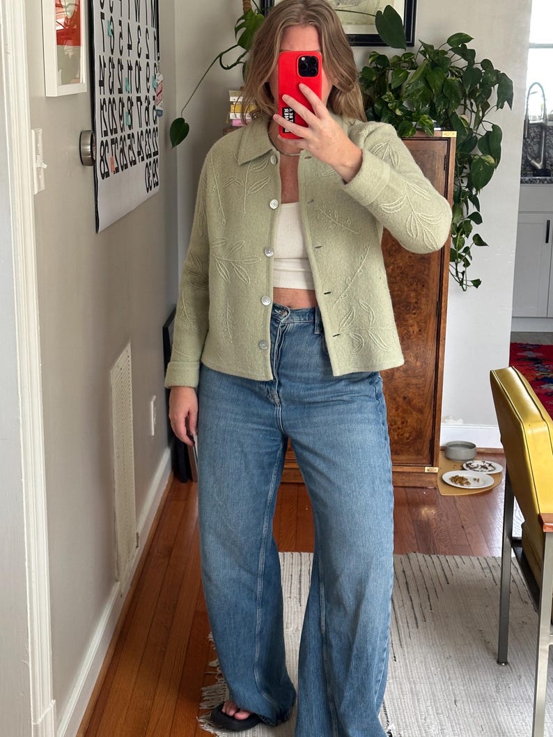 May include: A light green jacket with a leaf pattern, paired with blue jeans. The jacket has a button-up front and a collar. The jeans are a light wash, wide-leg style. The outfit is completed with black sandals.