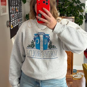 May include: A light gray sweatshirt with the text "NORTH CAROLINA" in a blue and white design. The sweatshirt also features the words "ATHLETIC CLUB" above the main text. The person in the image is wearing light blue jeans.