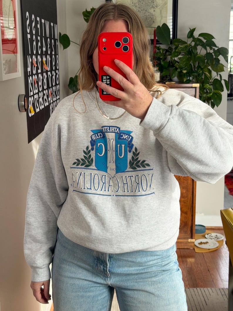 May include: A light gray sweatshirt with the text "NORTH CAROLINA" in a blue and white design. The sweatshirt also features the words "ATHLETIC CLUB" above the main text. The person in the image is wearing light blue jeans.