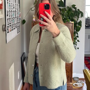 May include: A light green jacket with a leaf pattern. The jacket has a button-up front and a collared neckline. The sleeves are long, and the jacket appears to be made of a soft, textured fabric. The jacket is paired with blue jeans.
