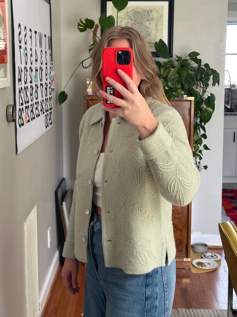 May include: A light green jacket with a leaf pattern. The jacket has a button-up front and a collared neckline. The sleeves are long, and the jacket appears to be made of a soft, textured fabric. The jacket is paired with blue jeans.