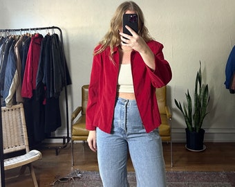 May include: A red corduroy jacket with a zip fastening and long sleeves. The jacket is worn over a white vest top and light blue jeans.