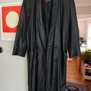 May include: A long, black leather coat with a notched collar and double-breasted button closure. The coat has two front pockets and is hanging on a wooden hanger. The coat is a classic style.