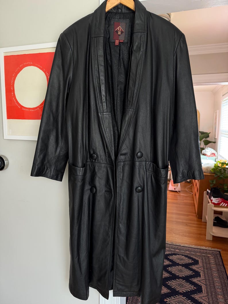 May include: A long, black leather coat with a notched collar and double-breasted button closure. The coat has two front pockets and is hanging on a wooden hanger. The coat is a classic style.
