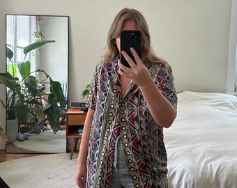 May include: A short-sleeved button-down shirt with a colorful geometric pattern in shades of red, blue, black, and beige. The shirt has a relaxed fit and a collar.