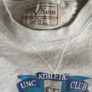 May include: A heather gray sweatshirt with a crew neck and a stitched V-neck detail. The sweatshirt features a patch with the text "UNC ATHLETIC CLUB" in blue and white. A tag reads "GALT SAND" and "MADE IN U.S.A."
