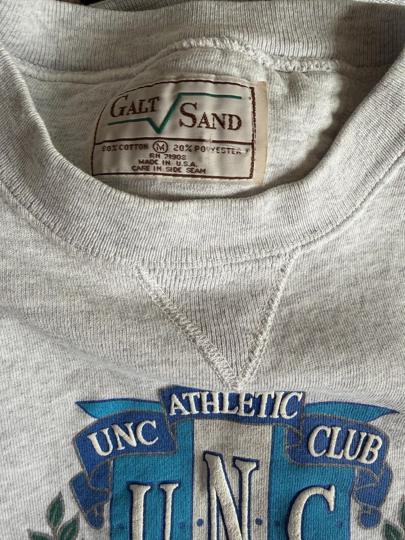 May include: A heather gray sweatshirt with a crew neck and a stitched V-neck detail. The sweatshirt features a patch with the text "UNC ATHLETIC CLUB" in blue and white. A tag reads "GALT SAND" and "MADE IN U.S.A."