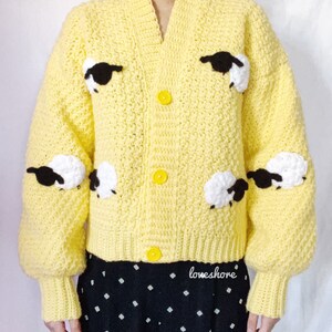 May include: A yellow crocheted cardigan with black and white sheep appliques. The cardigan has a V-neck and button closure. The sleeves are long and puffy. The cardigan is cropped and has a ribbed hem. The text "loveshore" is visible on the bottom of the cardigan.