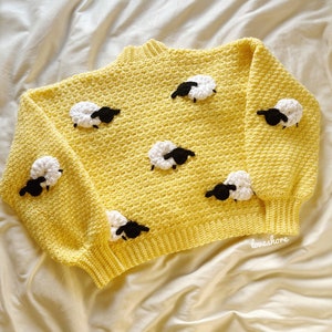 May include: A yellow crocheted sweater with a crew neck and a pattern of white sheep with black faces. The sweater has a relaxed fit and is perfect for a casual look.