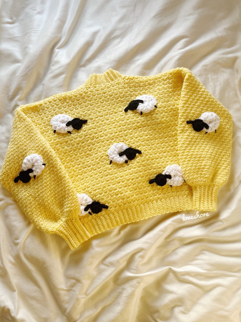 May include: A yellow crocheted sweater with a crew neck and a pattern of white sheep with black faces. The sweater has a relaxed fit and is perfect for a casual look.