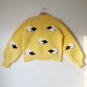 May include: Yellow crocheted sweater with black and white sheep appliques. The sweater has long sleeves and a round neckline.