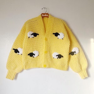 May include: A yellow crocheted cardigan with black and white sheep appliques. The cardigan has a v-neck and button closure.