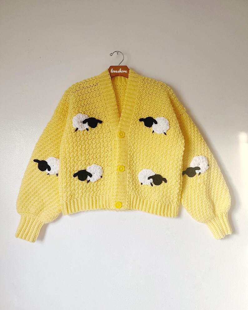 May include: A yellow crocheted cardigan with black and white sheep appliques. The cardigan has a v-neck and button closure.
