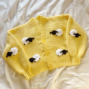 May include: A yellow crocheted cardigan with black and white crocheted sheep appliques. The cardigan has a button closure and is laid flat on a white surface.