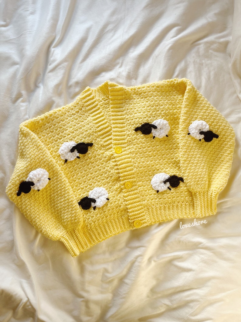 May include: A yellow crocheted cardigan with black and white crocheted sheep appliques. The cardigan has a button closure and is laid flat on a white surface.