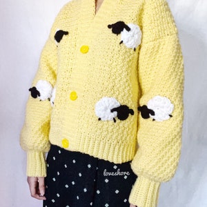 May include: A yellow crocheted cardigan with black and white sheep appliques. The cardigan has a V-neck and button closure. The sleeves are long and puffy. The cardigan is worn over a black and white polka dot skirt. The text "loveshore" is visible on the skirt.