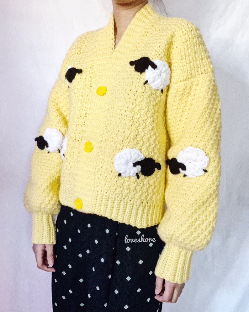 May include: A yellow crocheted cardigan with black and white sheep appliques. The cardigan has a V-neck and button closure. The sleeves are long and puffy. The cardigan is worn over a black and white polka dot skirt. The text "loveshore" is visible on the skirt.