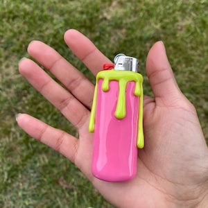 May include: A pink lighter with a lime green dripping design. The lighter has a silver metal top with a red button. The lighter is held in a person's hand against a background of green grass.