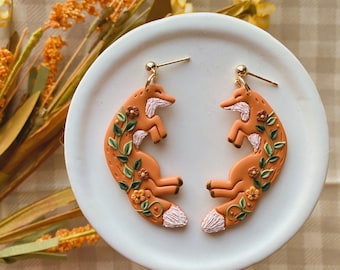 May include: A pair of orange fox earrings with floral details. The foxes are curled up with their tails wrapped around them. The earrings are made of polymer clay and have gold hooks.