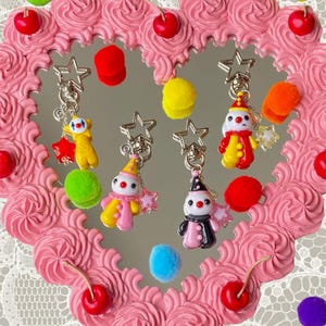 Peut inclure: Trois porte-clés en forme de clown avec des fermoirs en forme d'étoile. Un clown est jaune et rouge, un est jaune et rose, et un est noir et rose. Chacun comporte une petite breloque étoile et est présenté sur un cadre de miroir en forme de cœur décoré de tourbillons de glaçage rose, de cerises rouges et de pompons colorés.