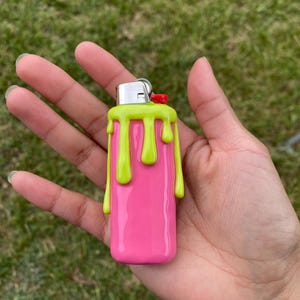 Puede incluir: Un encendedor rosa con un dise&ntilde;o de pintura goteante verde lima. El encendedor tiene una parte superior de metal plateado con un bot&oacute;n rojo. El encendedor se sostiene en una mano sobre un fondo de hierba verde.