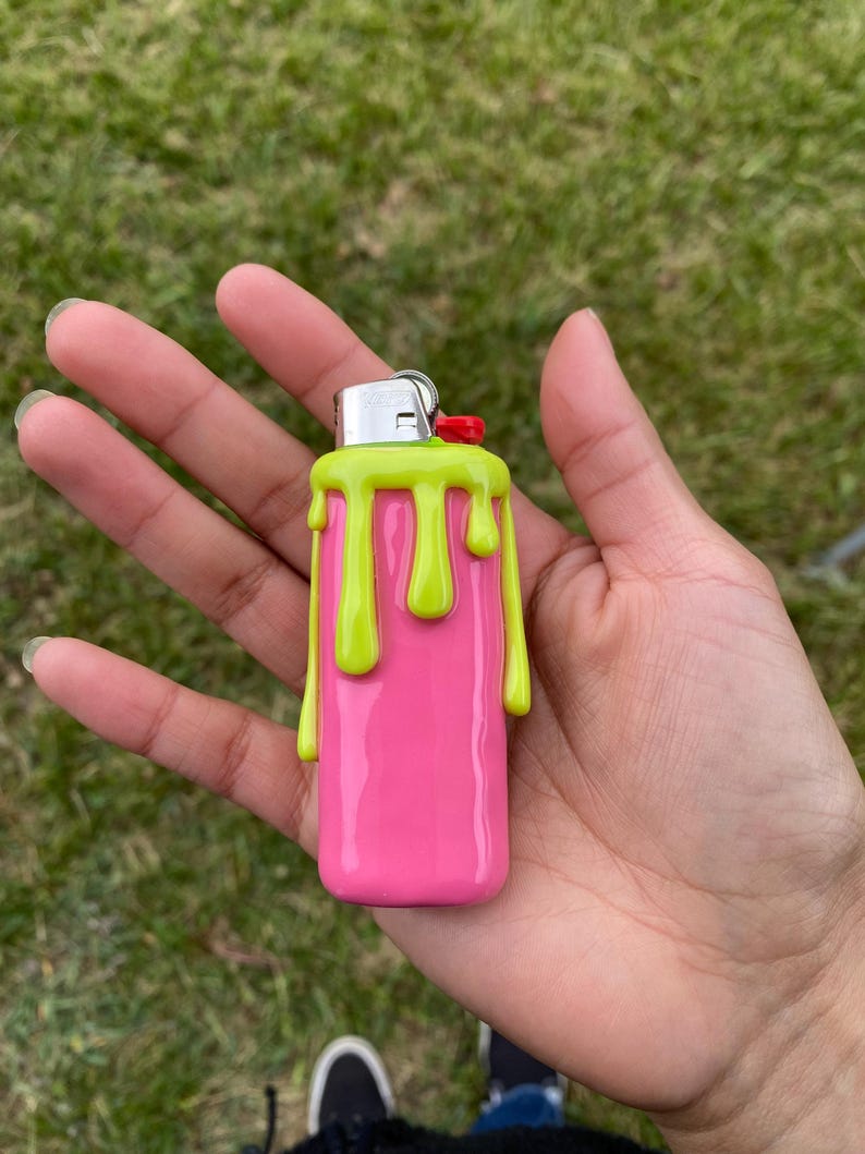 Puede incluir: Un encendedor rosa con un dise&ntilde;o de pintura goteante verde lima. El encendedor tiene una parte superior de metal plateado con un bot&oacute;n rojo. El encendedor se sostiene en una mano sobre un fondo de hierba verde.
