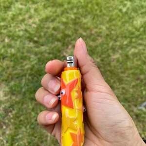May include: A yellow and orange glass lighter with a star design. The lighter has a metal top and two small glass accents. The lighter is held in a hand against a background of green grass.