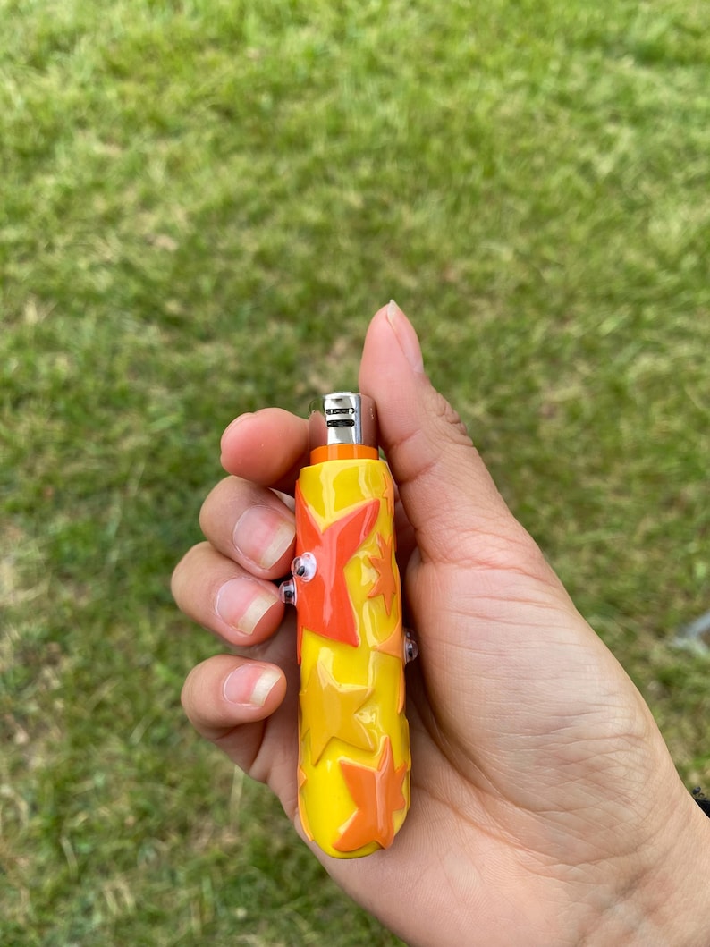 May include: A yellow and orange glass lighter with a star design. The lighter has a metal top and two small glass accents. The lighter is held in a hand against a background of green grass.