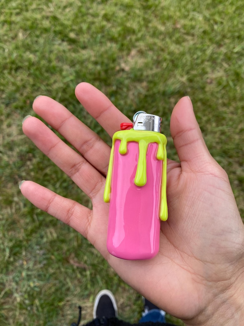May include: A pink lighter with a lime green dripping design. The lighter has a silver metal top with a red button. The lighter is held in a person's hand against a background of green grass.