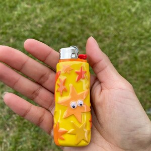 May include: A yellow lighter decorated with orange starfish and googly eyes. The lighter has a red button and a silver metal top. The background is green grass.