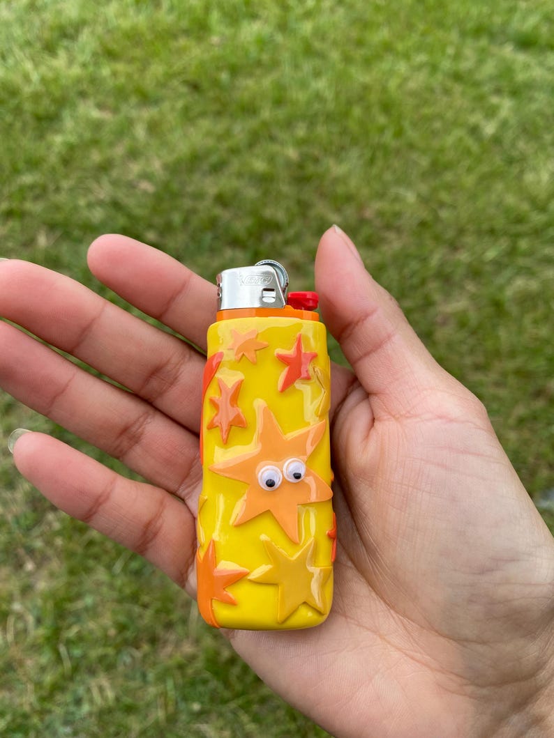 May include: A yellow lighter decorated with orange starfish and googly eyes. The lighter has a red button and a silver metal top. The background is green grass.
