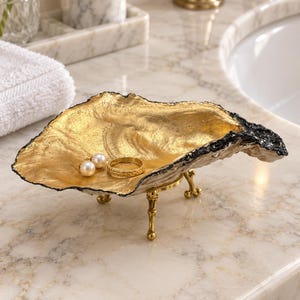 May include: A decorative oyster shell dish featuring a metallic gold interior and a dark, textured exterior. The shell is supported by three small, slender gold-toned metal legs and contains a gold ring and two pearl stud earrings.