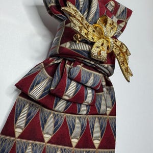 May include: A burgundy tie with a repeating geometric pattern in blue and white, secured with a gold-colored bow-shaped brooch. The tie is knotted, showcasing the pattern and the brooch's intricate design. The brooch is adorned with small, sparkling details.