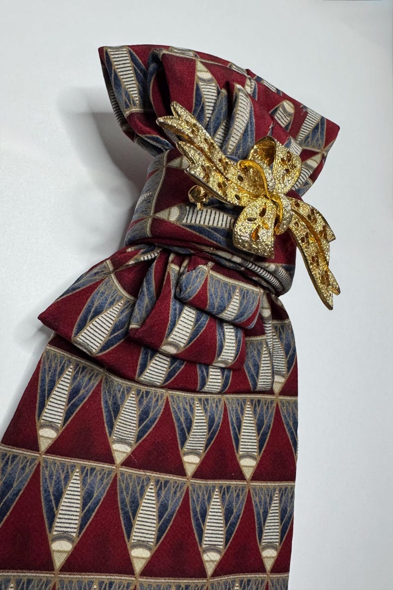 May include: A burgundy tie with a repeating geometric pattern in blue and white, secured with a gold-colored bow-shaped brooch. The tie is knotted, showcasing the pattern and the brooch's intricate design. The brooch is adorned with small, sparkling details.
