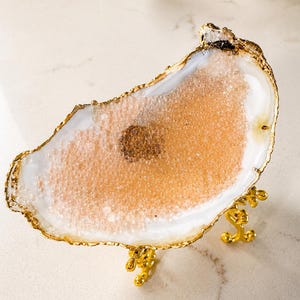 Op de afbeelding: Een decoratieve schaal gemaakt van een witte en doorschijnende agaatplak met een natuurlijke, onregelmatige rand. De rand is afgewerkt in metallic goud en de schaal wordt ondersteund door drie sierlijke, goudkleurige metalen poten.