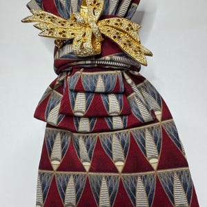 May include: A patterned necktie in shades of burgundy, blue, and beige, adorned with a gold-colored bow-shaped accessory. The tie features a repeating geometric design. The bow is embellished with small, sparkling details.
