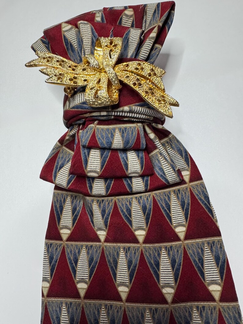 May include: A patterned necktie in shades of burgundy, blue, and beige, adorned with a gold-colored bow-shaped accessory. The tie features a repeating geometric design. The bow is embellished with small, sparkling details.