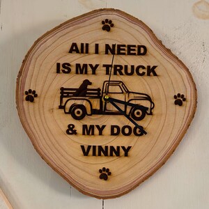 May include: A wooden clock with a rustic woodgrain finish. The clock face features the text "All I need is my truck & my dog Vinny" with a silhouette of a truck and a dog. The clock is decorated with paw prints.