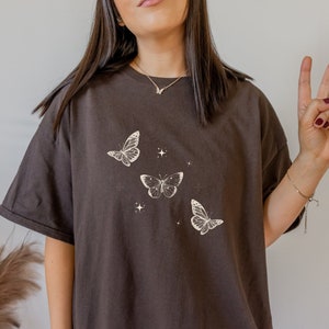 Könnte beinhalten: Ein braunes Oversize-T-Shirt mit einem Motiv aus drei weißen Schmetterlingen und Sternen.