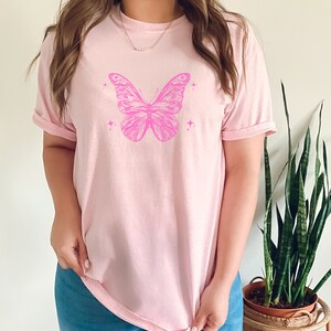 May include: A pink t-shirt featuring a pink butterfly graphic with small stars. The butterfly has detailed wings and a body.
