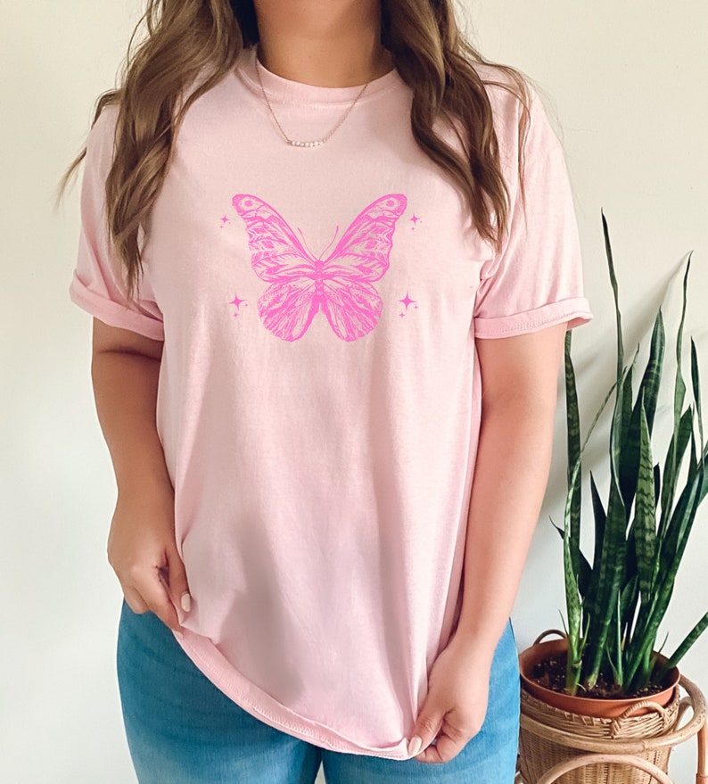 May include: A pink t-shirt featuring a pink butterfly graphic with small stars. The butterfly has detailed wings and a body.