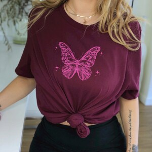 May include: A maroon t-shirt featuring a pink butterfly graphic and a gold chain necklace with a cross pendant.