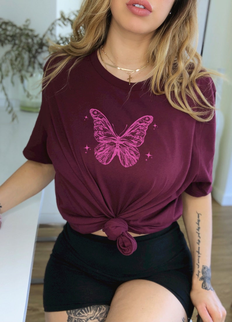 May include: A maroon t-shirt featuring a pink butterfly graphic and a gold chain necklace with a cross pendant.