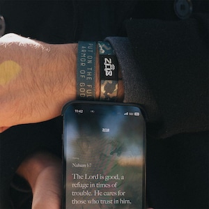 May include: A wrist adorned with two fabric bracelets and a smartphone. One bracelet is navy blue with the text "PUT ON THE FUL ARMOR OF GOD". The other is camouflage with the text "ZAS". The phone displays a Bible verse from Nahum 1:7.