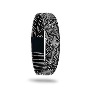 May include: Black and white patterned wristband with a geometric design. The wristband features a repeating pattern of lines, triangles, and dots. The band has a black velcro closure.
