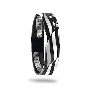 May include: Black and white elastic wristband featuring a design that resembles the American flag. The wristband has the text "WE NEVER WALK ALON" printed on it. The wristband is made of a stretchy material and has a black velcro closure.