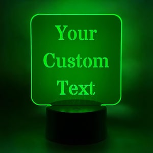 May include: A green LED night light with the words "Your Custom Text" illuminated on a square panel. The light sits on a black cylindrical base, creating a glowing effect in a dark setting.